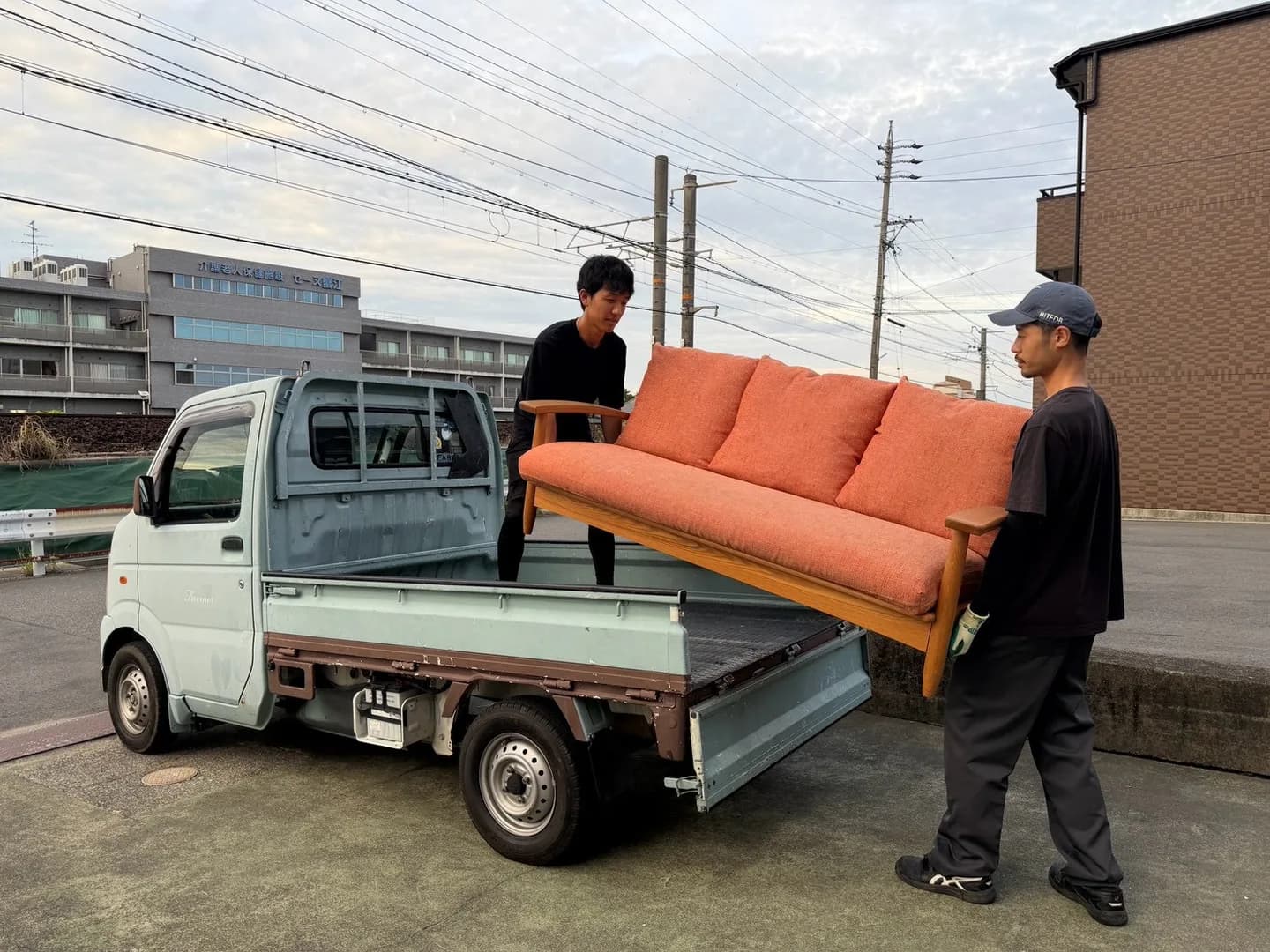 2人のスタッフがオレンジ色の木枠ソファを軽トラックの荷台付近で支える様子