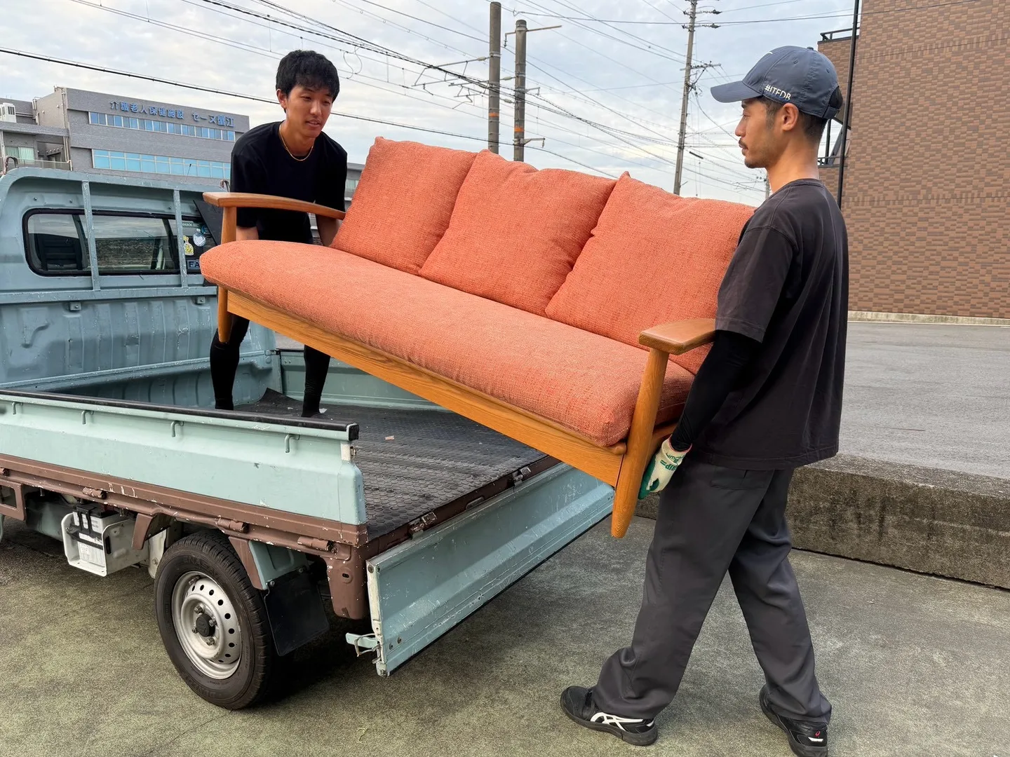 木製の肘掛け・脚・座面が見えるオレンジ色のソファをスタッフが支えている作業写真